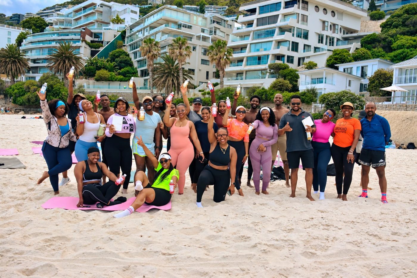 A big group of smiling people on a beach, holding up colourful bottled drinks and cheering at a Sober Woke sober bar event