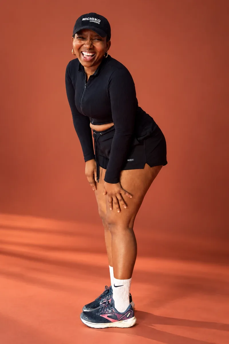 Thandokazi Ntshinga laughing out loud, leaning forward in a black cap and outfit against a terracotta backdrop
